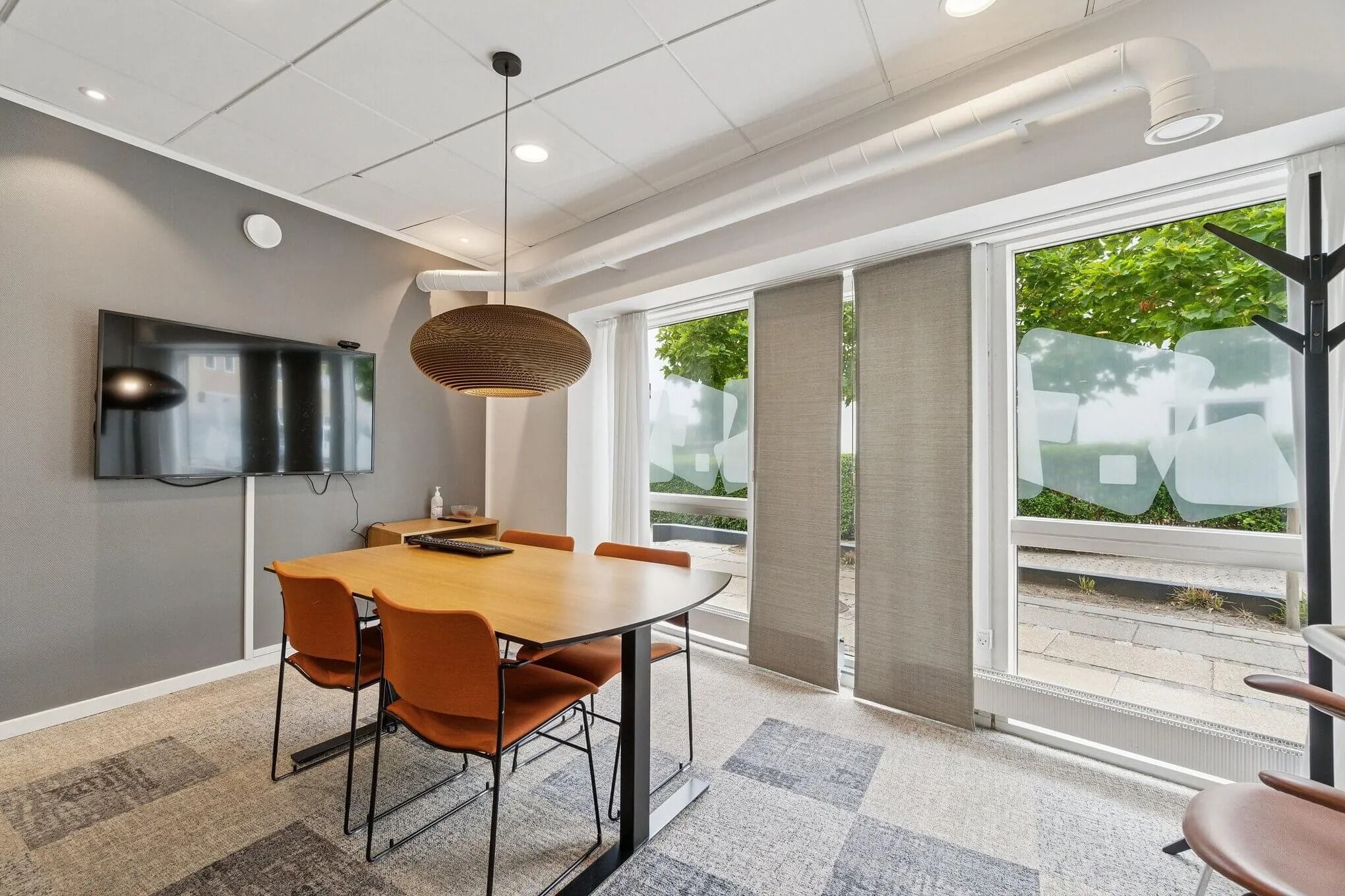 Velholdt mødelokale med bord, stole og tv. Fladt loft med hvide loftbelægninger og lysekroner. Moderne møbler og en stor