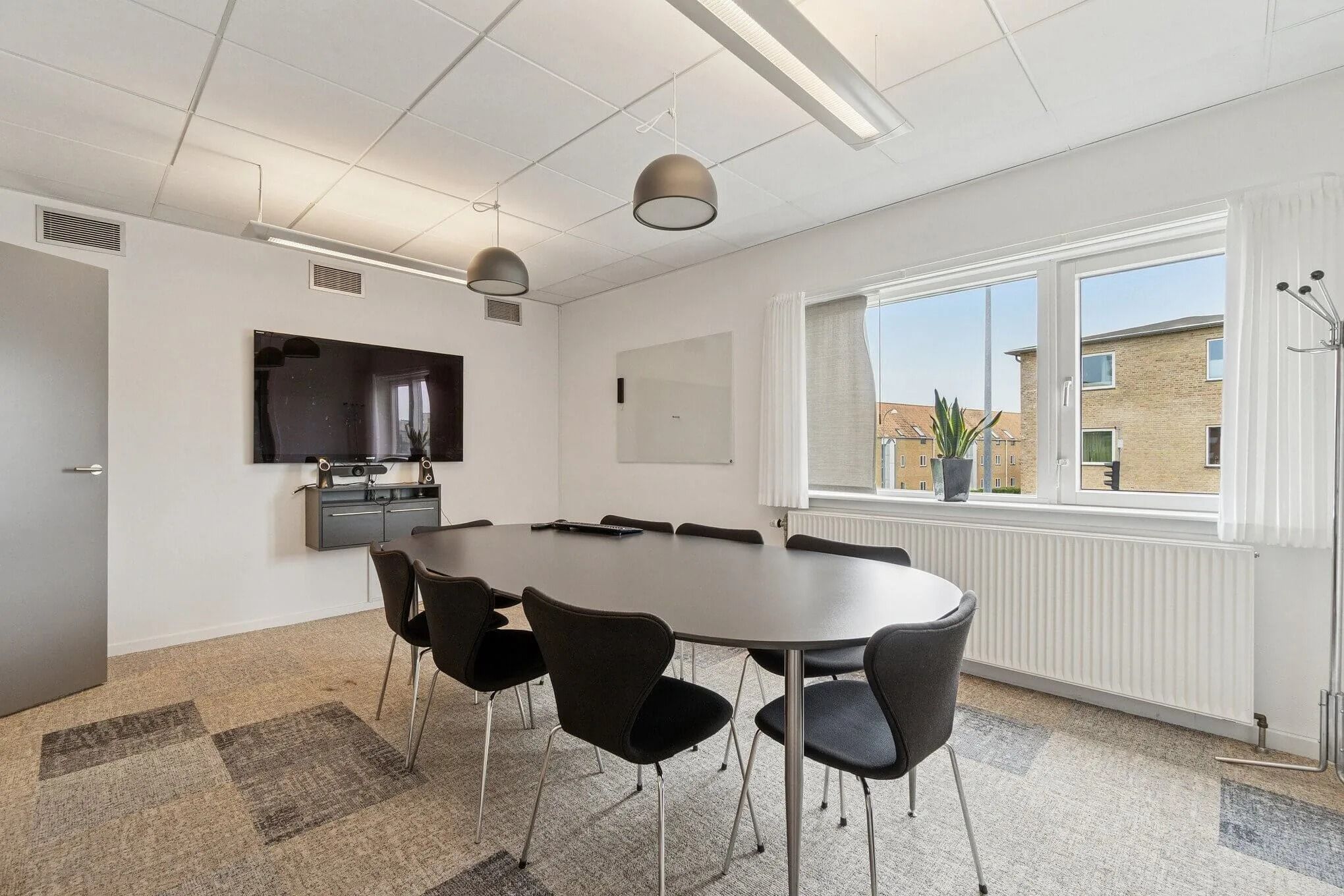 Inde i et mødelokale er der en stor ovalt bord, otte stole og en skærm samt en plante på vindueskarmen