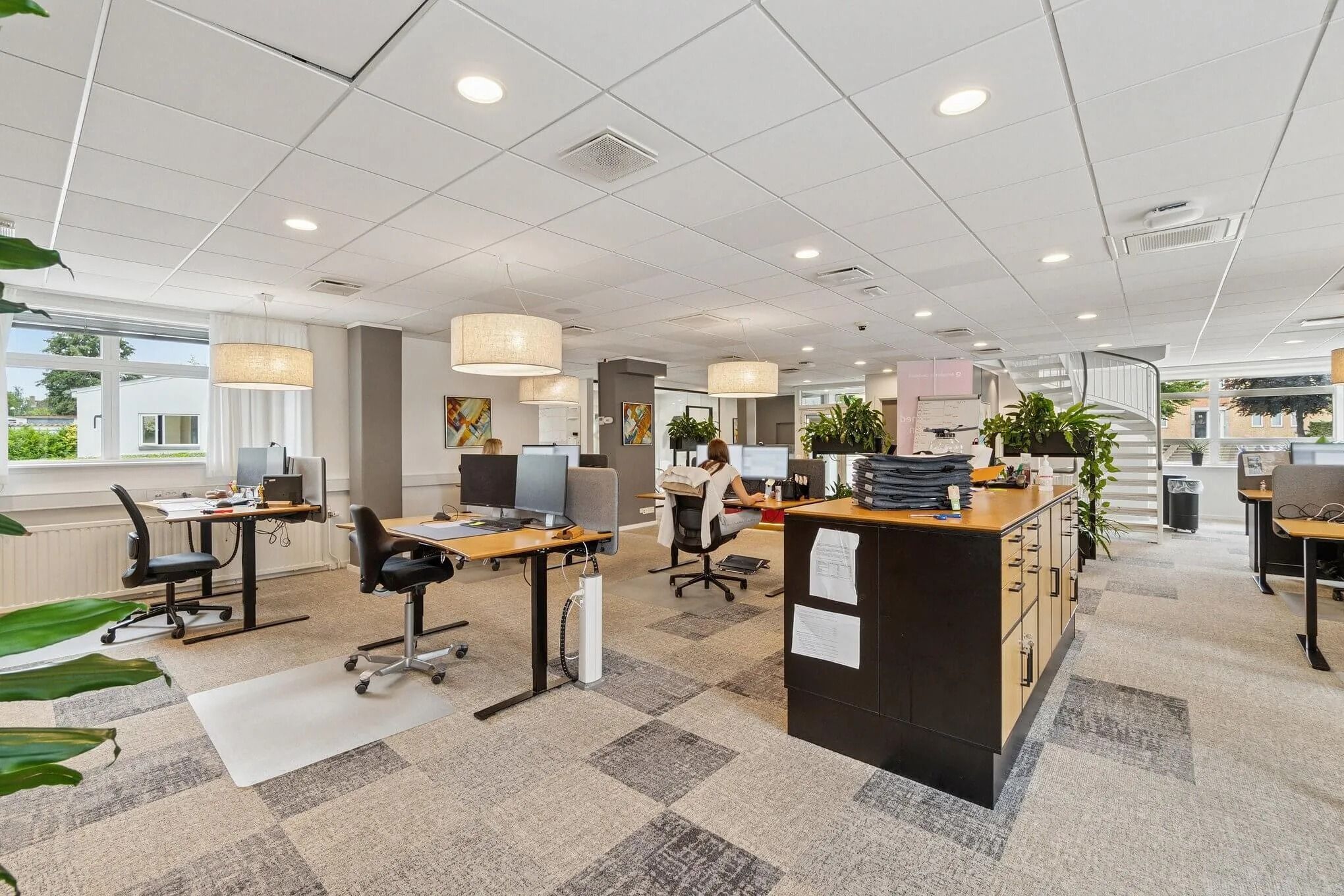 Kontorområde med moderne møbler og store vinduer for lyst arbejdsmiljø. Styrket fokus på grønne planter for bedre atmosfære.