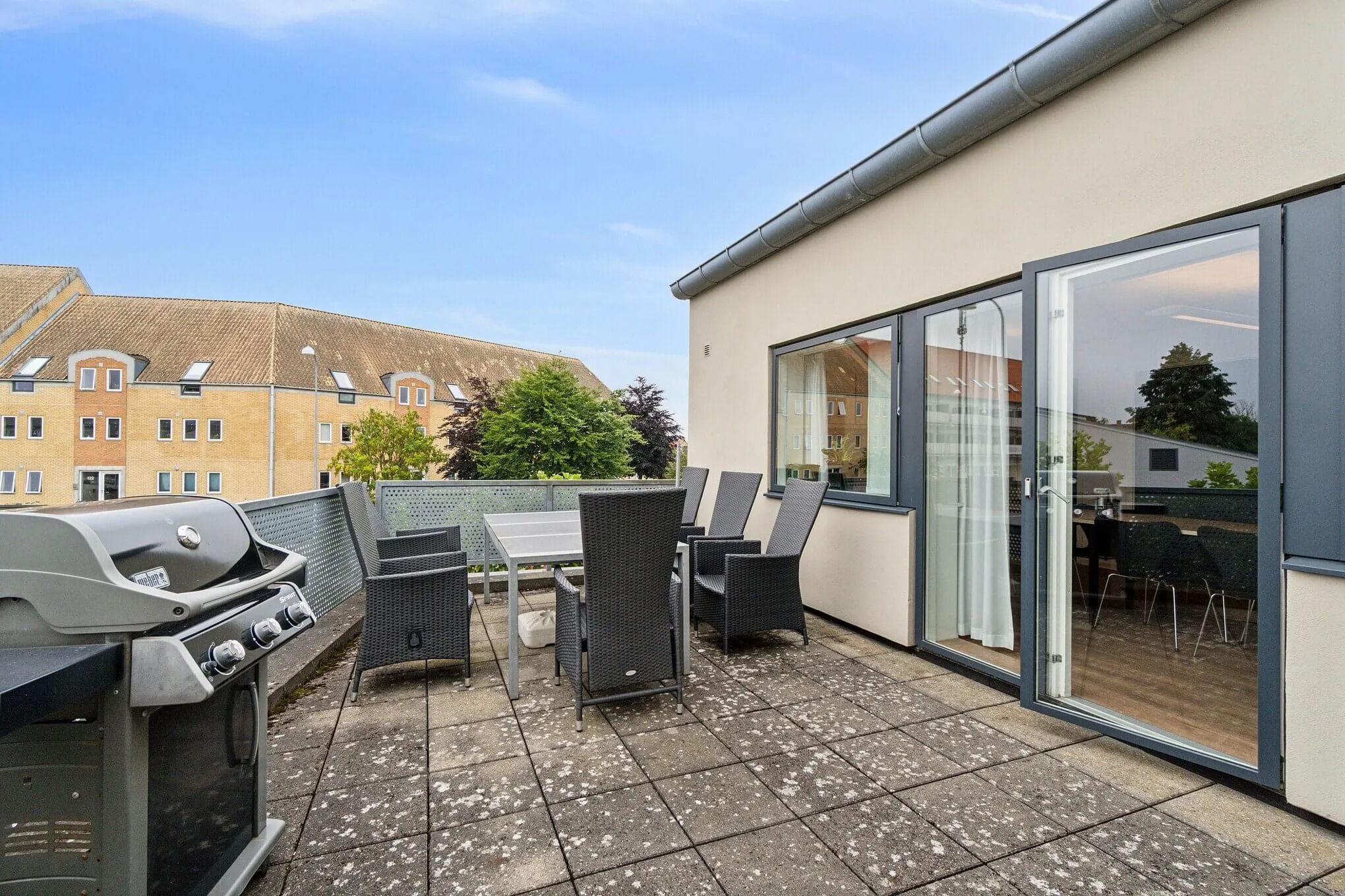 Terrasse med grill, møbler og udsigt til et nærliggende bygningskompleks med lyse facader og en solrig himmel