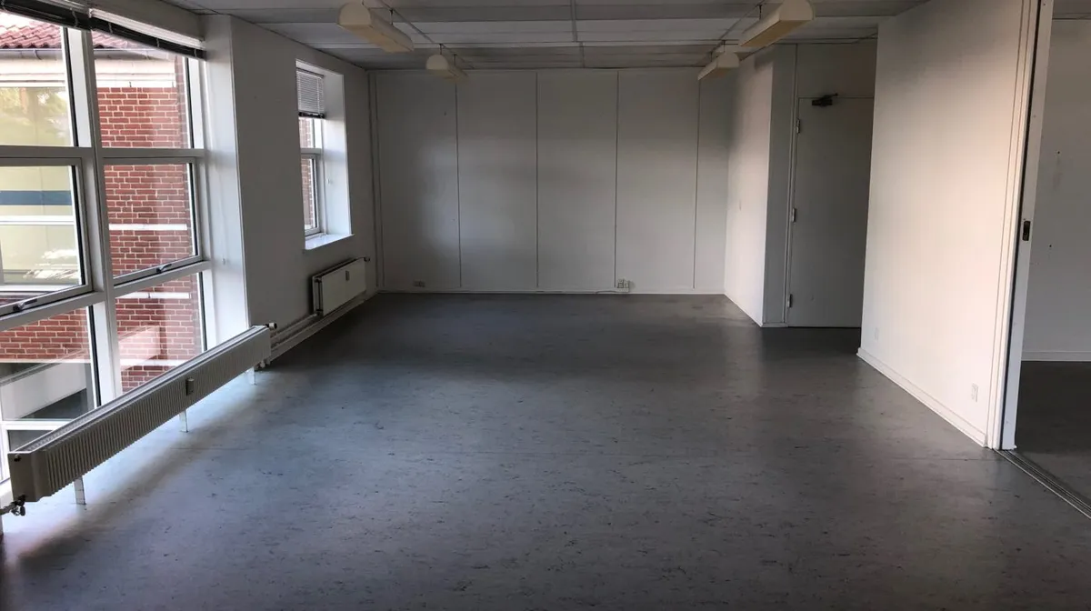 En stor lys og indbydende kontorlokal med hvide vægge og gulv, store vinduer og en åben dør, samt flere radiatorer og
