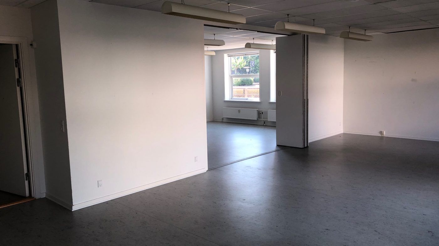 Et stort, lyst og rummeligt lokale med lyse loftslampe, to vinduer og en hvid væg. Gulvet er lavet af lysegrå granit og