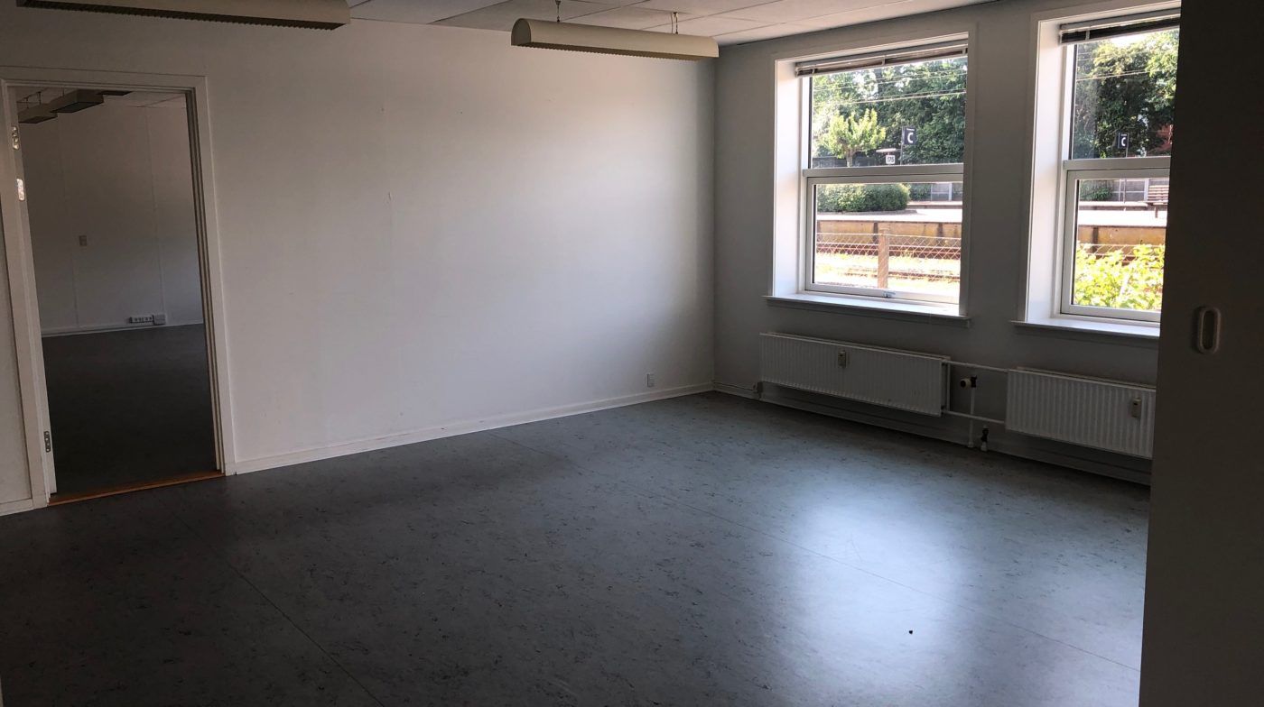 Et stort kontorlokale med hvide vægge og lysegulve med to store vinduer og radiatorer og to lange lysekroner