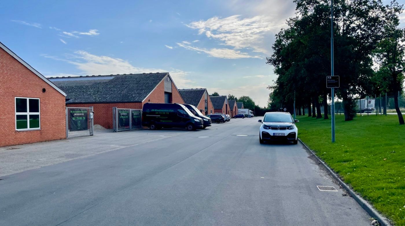 Et lille erhvervspark med fem værksteder og lagerlokaler. Parkerede biler og to busser udenfor. En bil kører langs en