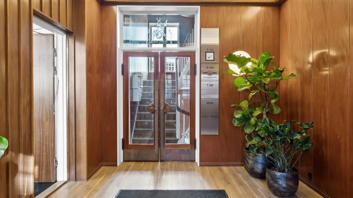 En entre med træpaneler og glasdøre, to store planter og elevatorpaneler med knapper til sal 1, 2 og 3