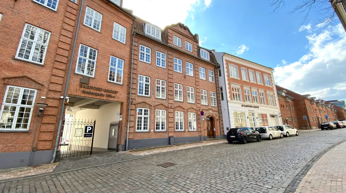Gade med gamle, røde murstensbygninger med mange vinduer og en parkeringsplads med biler foran Barfoed Group Nedergade 35b