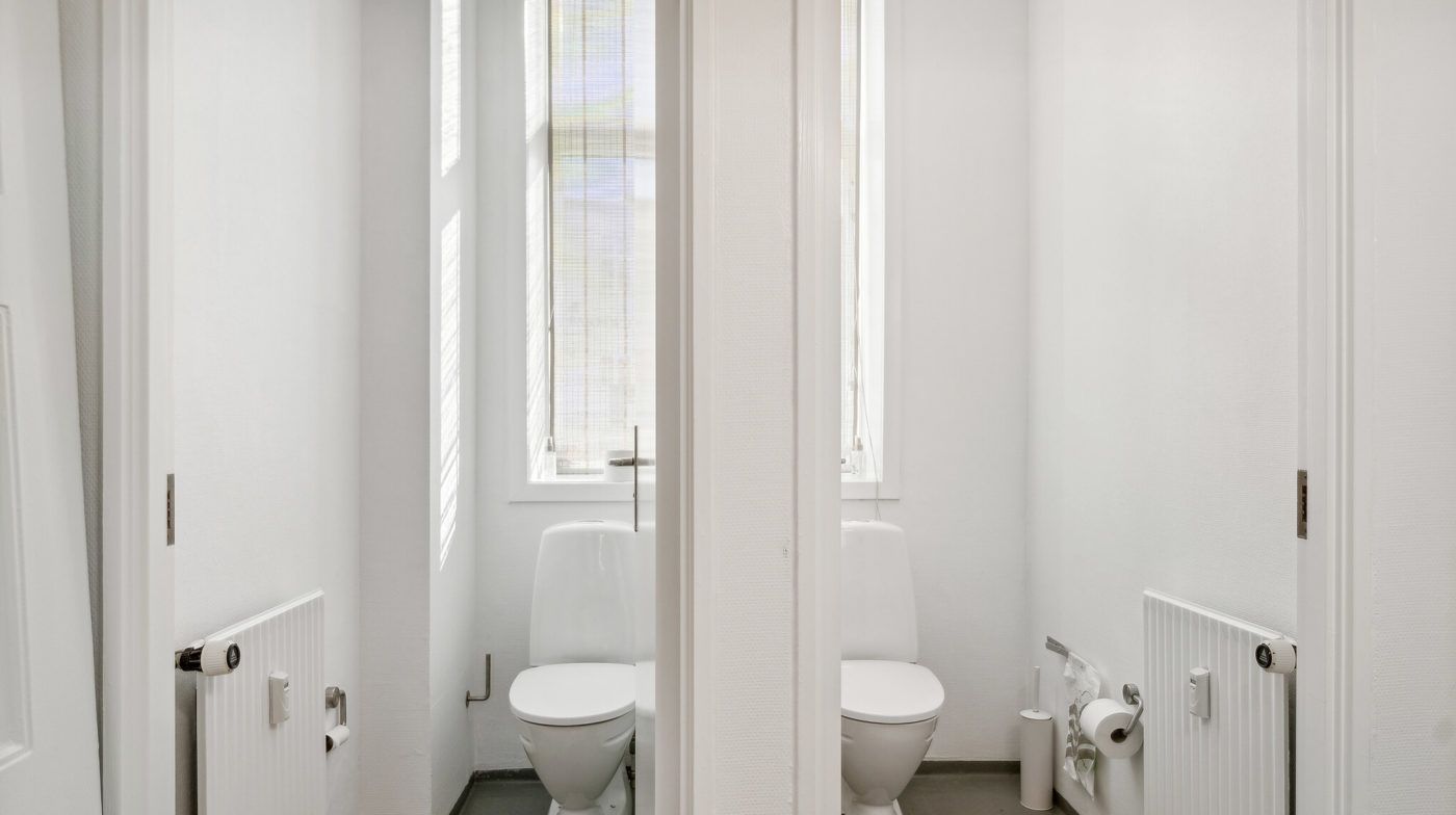 To toiletrum med hvide toiletter og radiatorer samt to toiletpapirruller på toiletvæggene