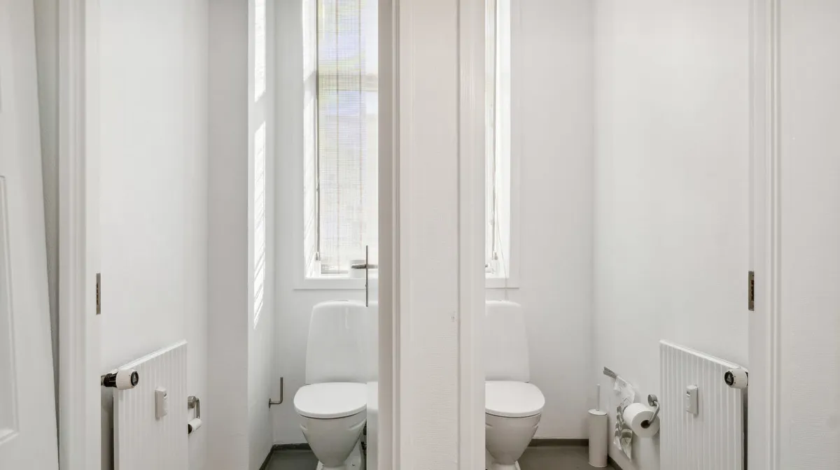 To toiletrum med hvide toiletter og radiatorer samt to toiletpapirruller på toiletvæggene