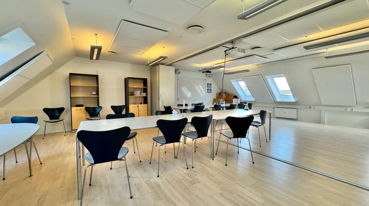 Kontor med to lange borde og flere stole, hvor det er lyse og tæt på vinduer