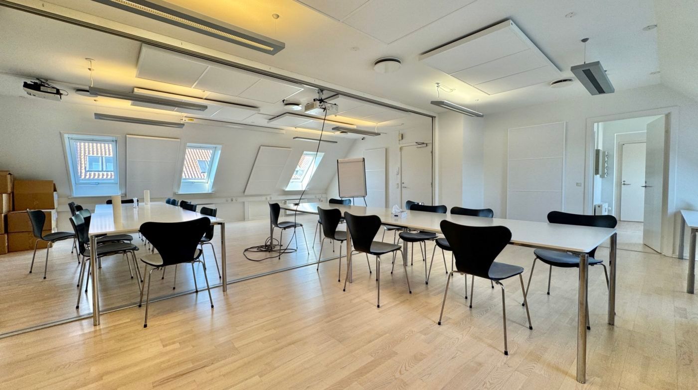 Kontorlokal med lydisolering, hvide vægge, trægulv og flere borde med stole samt en hvid tavle og et projektor