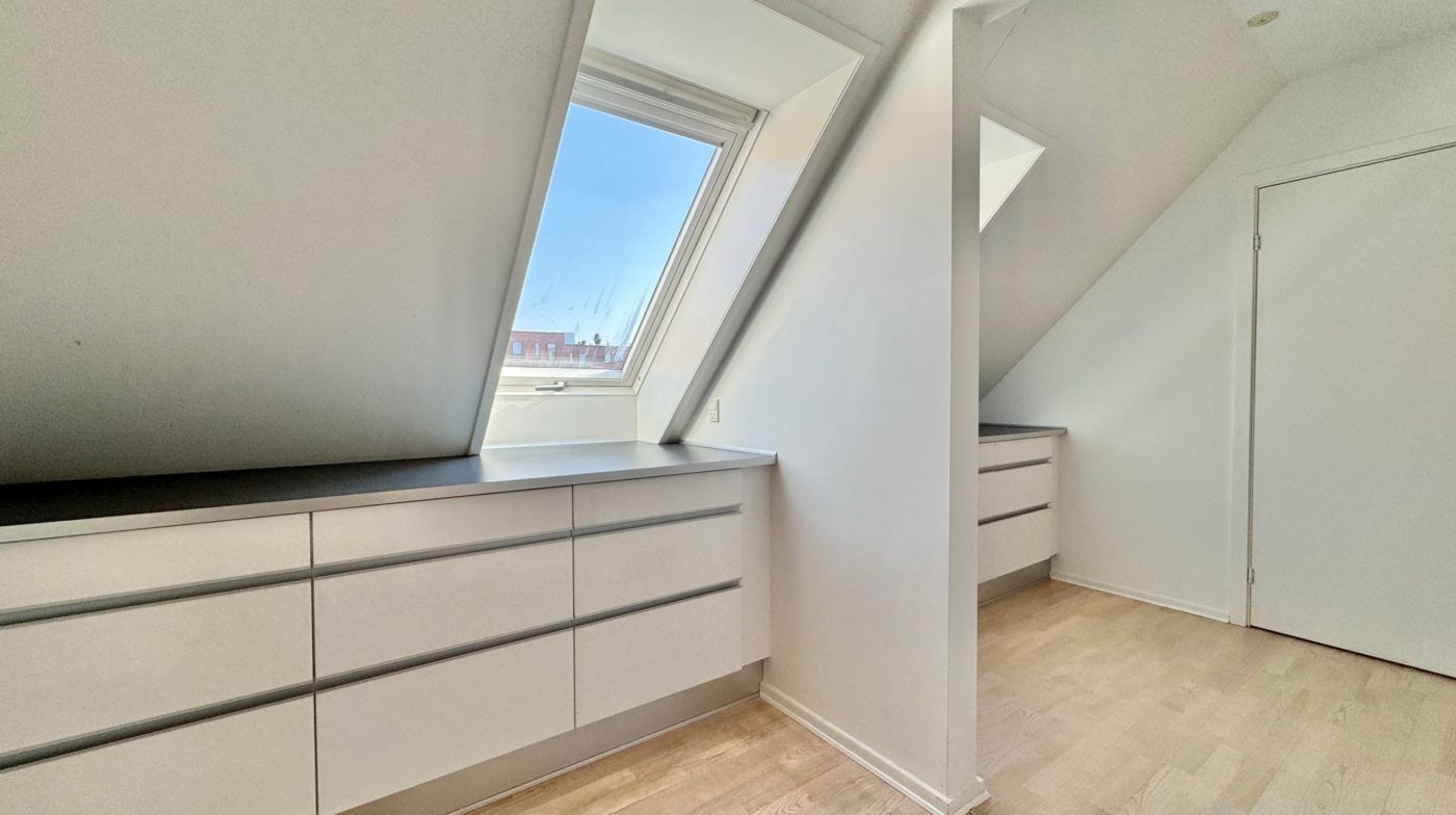 Et lille og rent loftsværelse med hvide vægge og loft, to trægulve, et vindue og en dør, der fører til et andet værelse