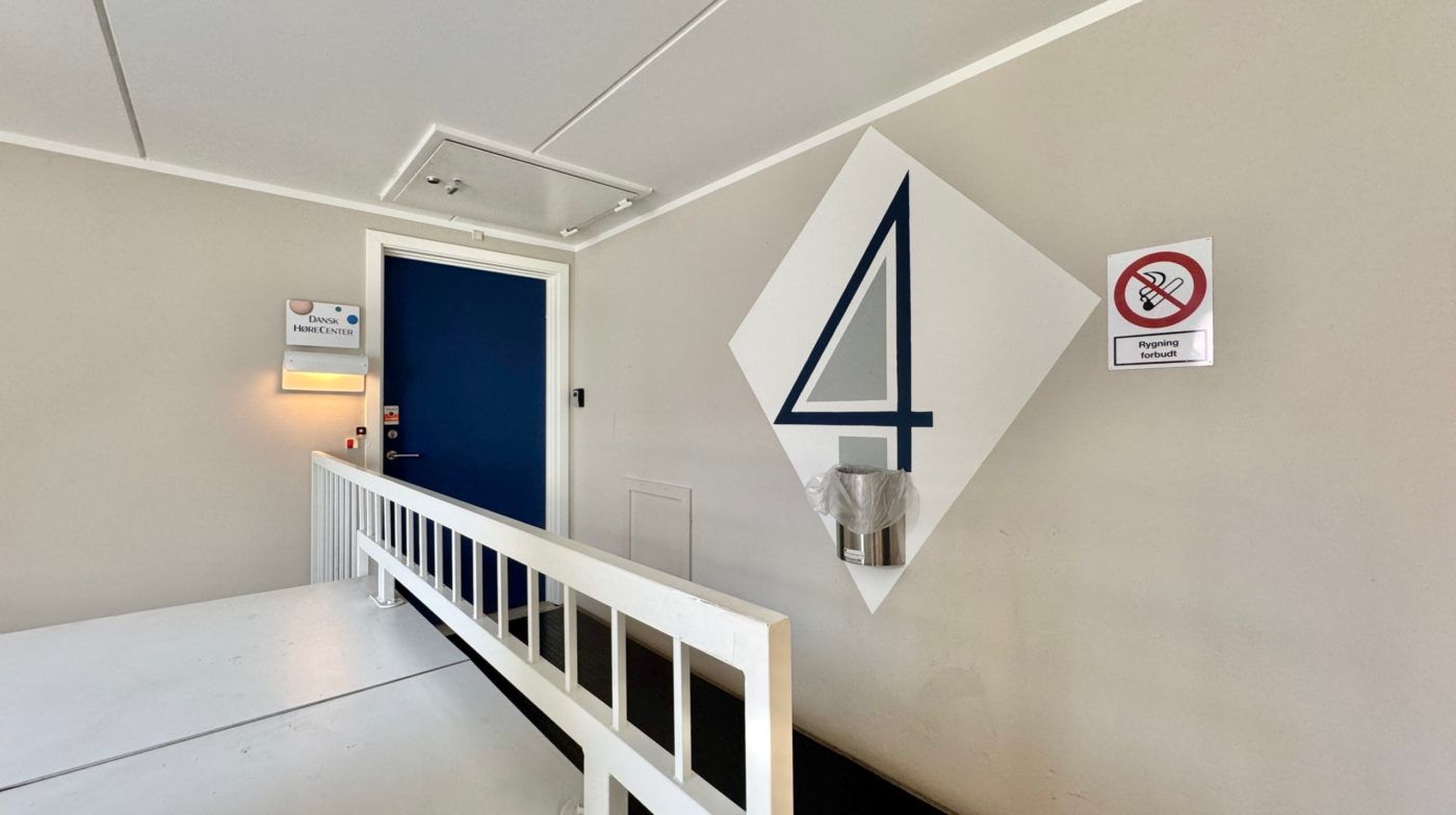 Stående skrånende trappeopgang med hvidt rækværk og hvid gulvbelægning. Til højre for trappen er der en stor vægmonteret