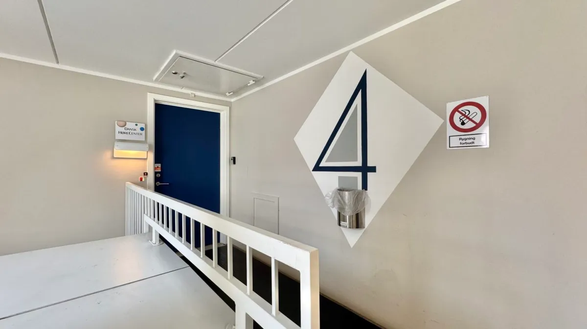 Stående skrånende trappeopgang med hvidt rækværk og hvid gulvbelægning. Til højre for trappen er der en stor vægmonteret