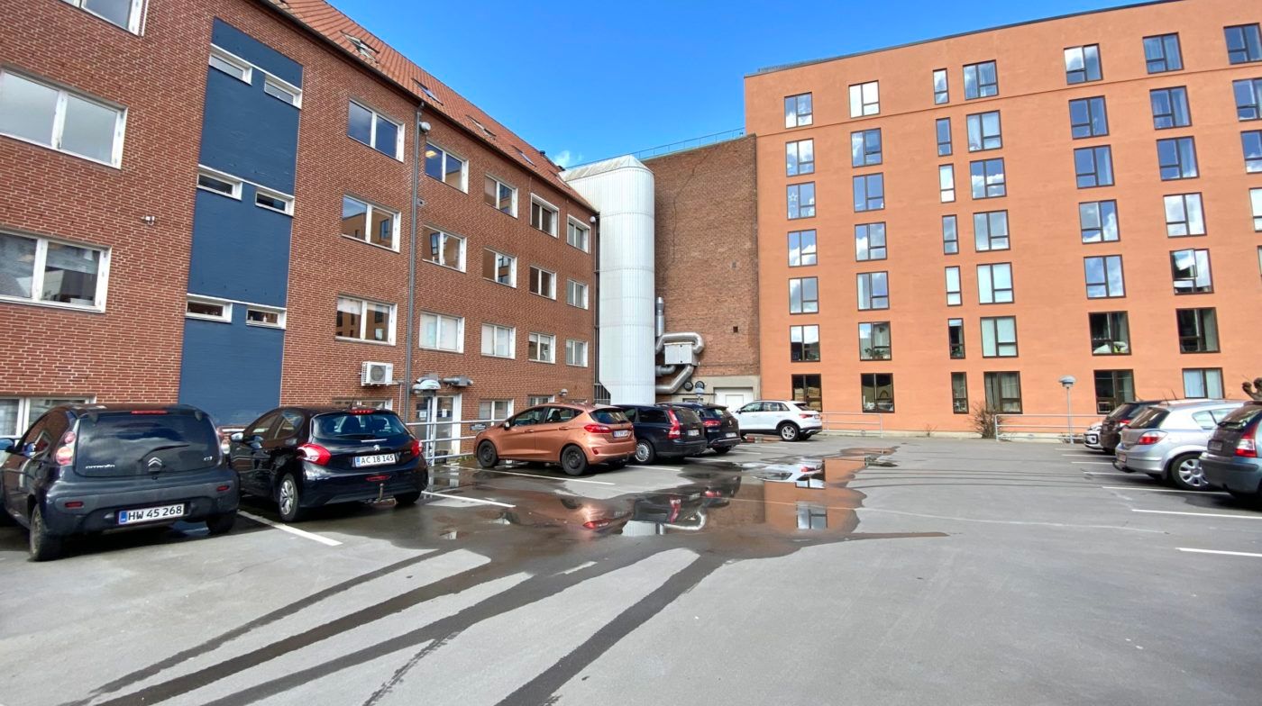 Flere biler parkeret i en parkeringsplads med to multifamilieboliger i baggrunden, og et hav af skyer i himlen