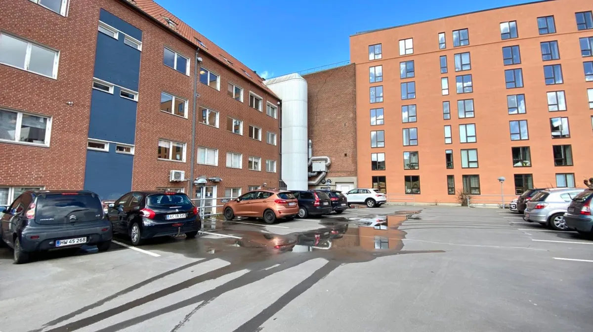 Flere biler parkeret i en parkeringsplads med to multifamilieboliger i baggrunden, og et hav af skyer i himlen