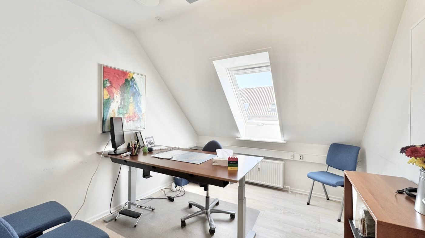 Et moderne kontorlokal med skrivebord, computer, billeder og blomster på et bord og en radiator under vinduet