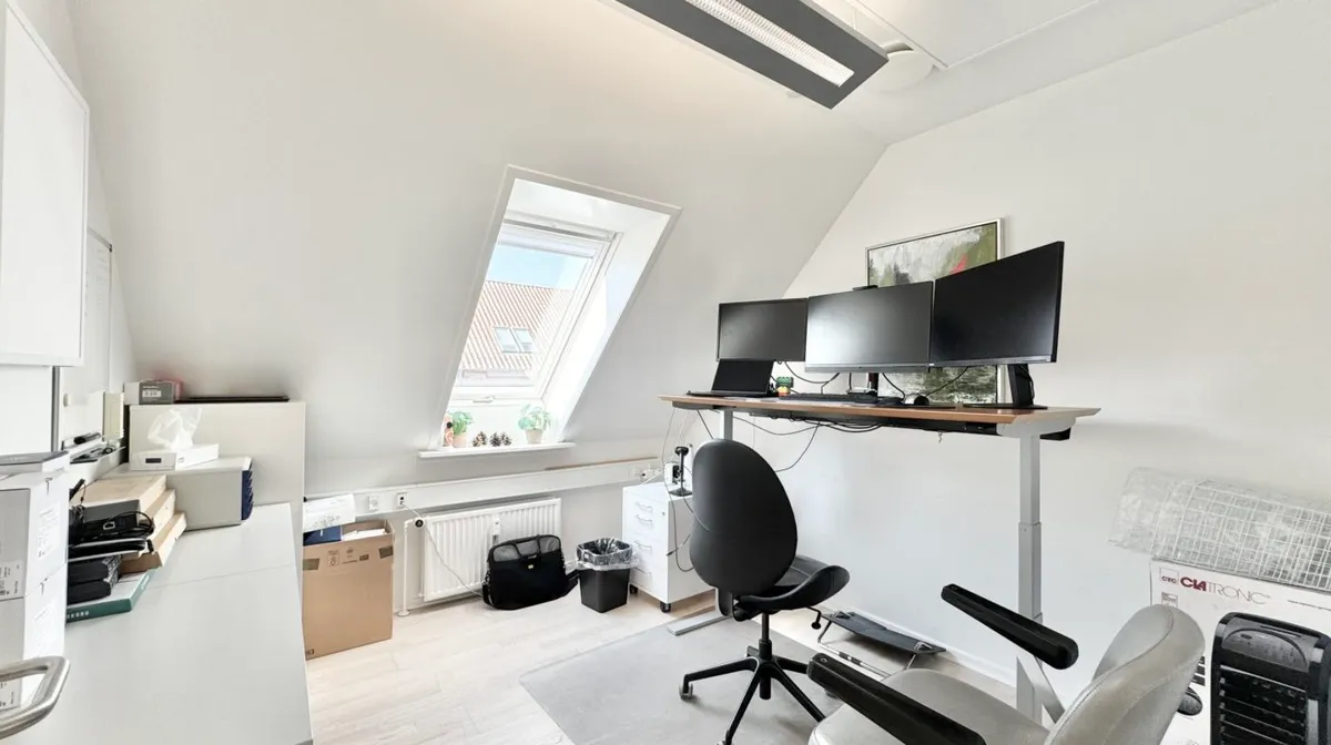 Et moderne kontorlokale med lyse vægge og loft samt skrivebord med computere og stol. Et vinduesrum med plante og lyse