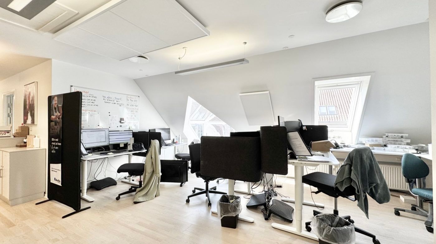 Kontorrum med to skriveborde og fire kontorstole i sort. Væggene er hvide og der er en skrivebordsvæg med fire computere og