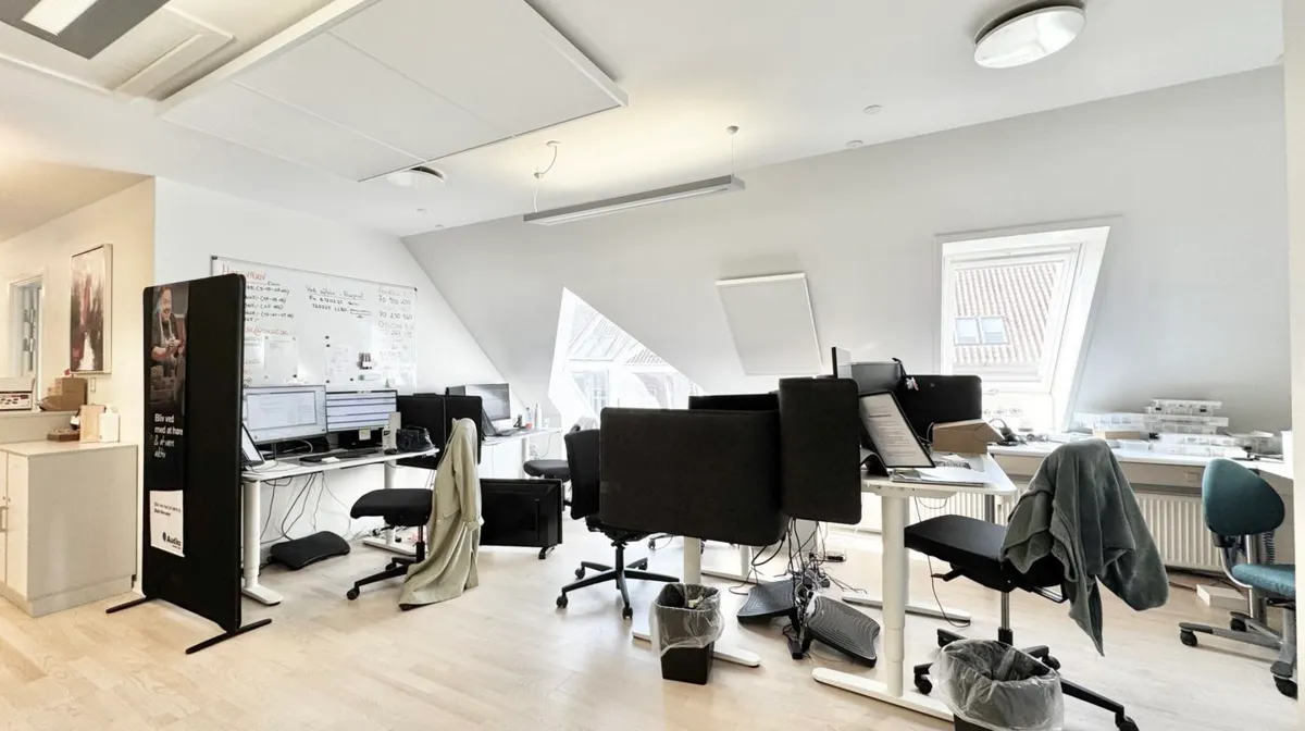 Kontorrum med to skriveborde og fire kontorstole i sort. Væggene er hvide og der er en skrivebordsvæg med fire computere og