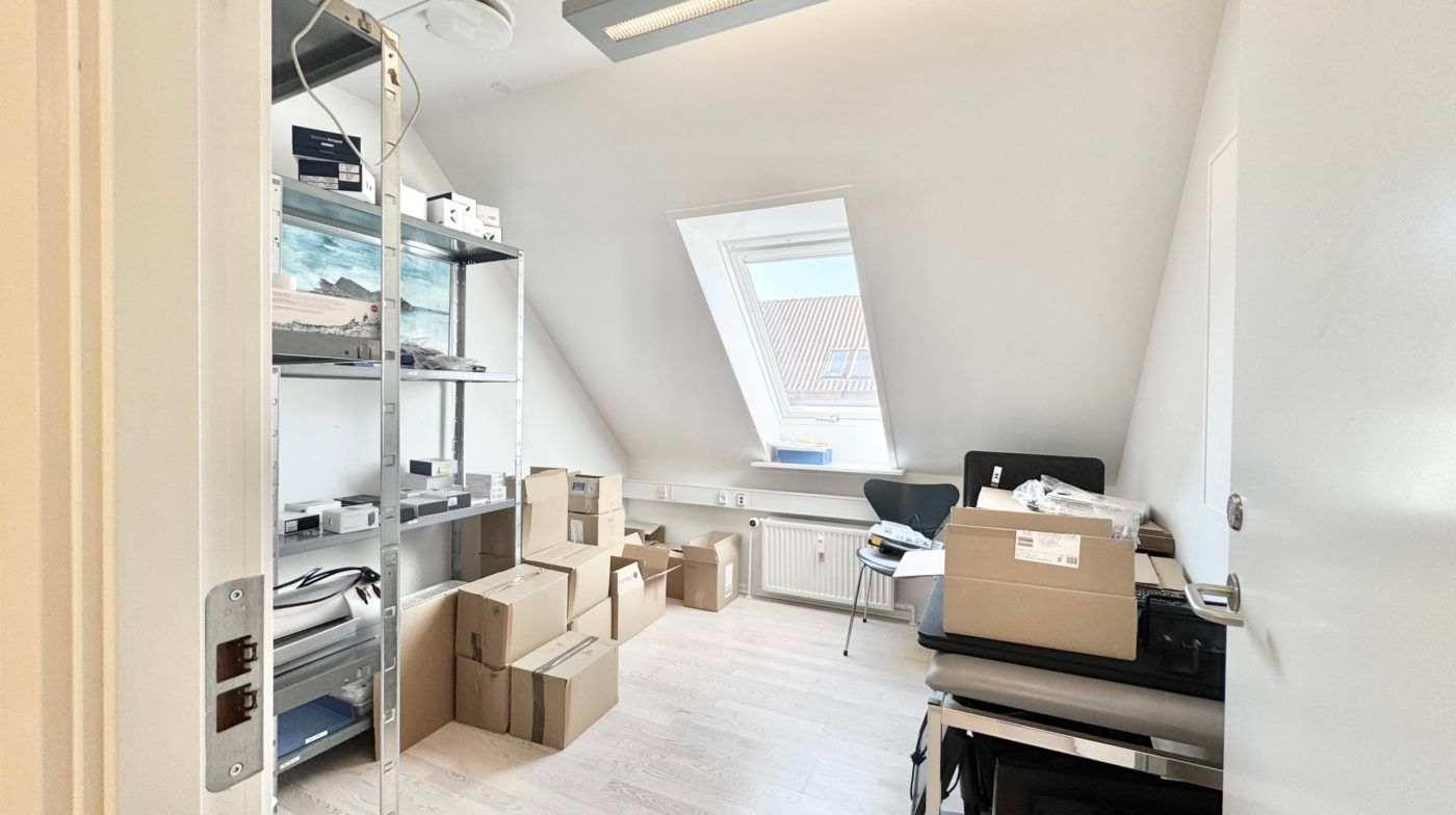 Et værelse med højt til loftet og vinduesindslag, flere kartoner og pakninger, samt højre hjørne med bord og stol.