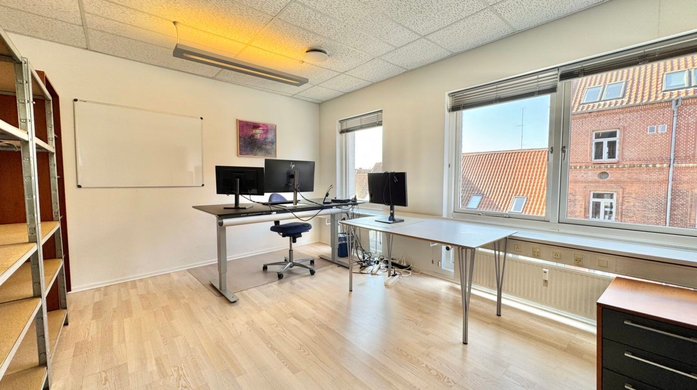 Kontorrum med lyse gulve, vinduer, skriveborde, stole og hvide vægge med en tavle og maleri.