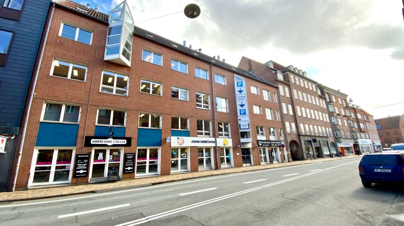 En bil kører forbi et bygningskompleks med flere butikker og kontorer. Bygningen har en blå facade og mange vinduer.