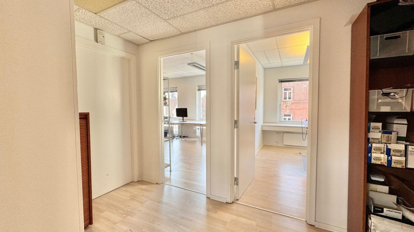 To kontordøre med indgang til et møbleret kontorlokale med en skrivebord, stol og en monitor samt et køkken med en brik.