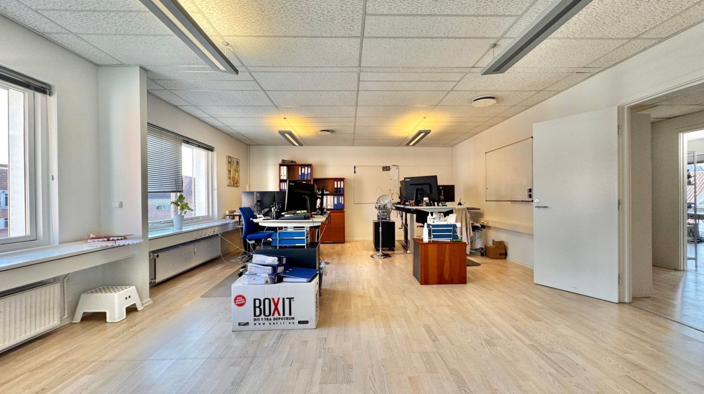 Kontor med trægulv, hvide vægge og to skriveborde med monitorer, kabinetter og plante.