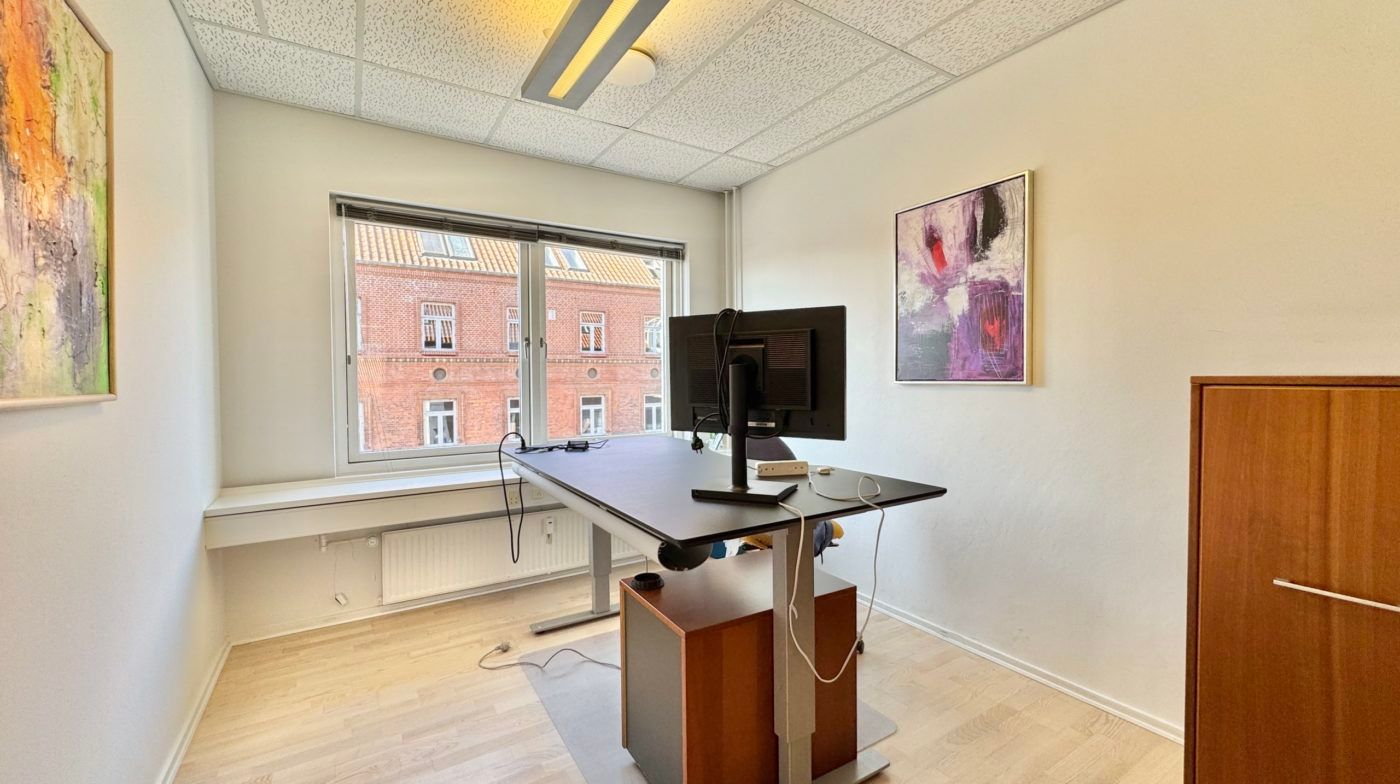 Kontorrum med en højdeskiftende skrivebord, computer, kabinet og malerier på væggen samt et vindue med udsigt til bygning