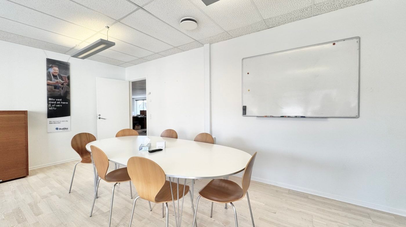Et mødelokale med hvide vægge og gulv samt runde borde og stole med høj ryglæn. En hvid tavle og en plakat hænger på væggen.