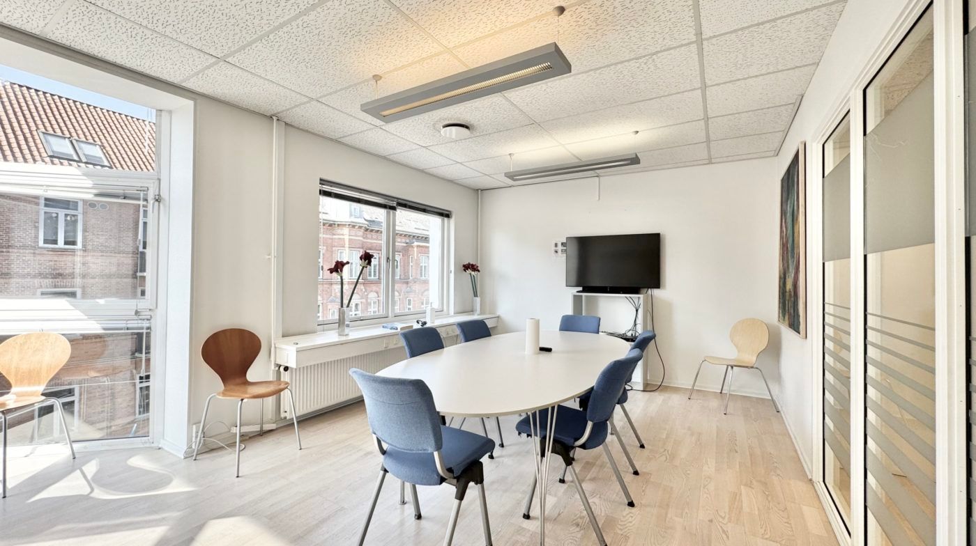 Et moderne mødelokale med en stor rund hvid bord og blå stole, samt en flatscreen tv med en højtalere på væggen.