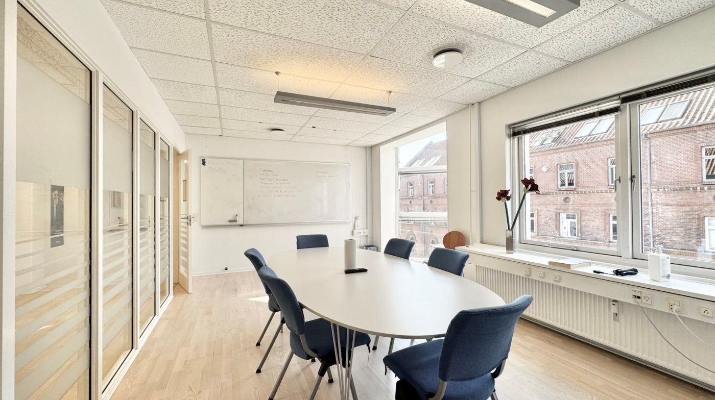 Et tomt konferencerum med hvide vægge, lyse lofter, et stort ovalt bord og blå stole samt en hvid tavle og vinduer med