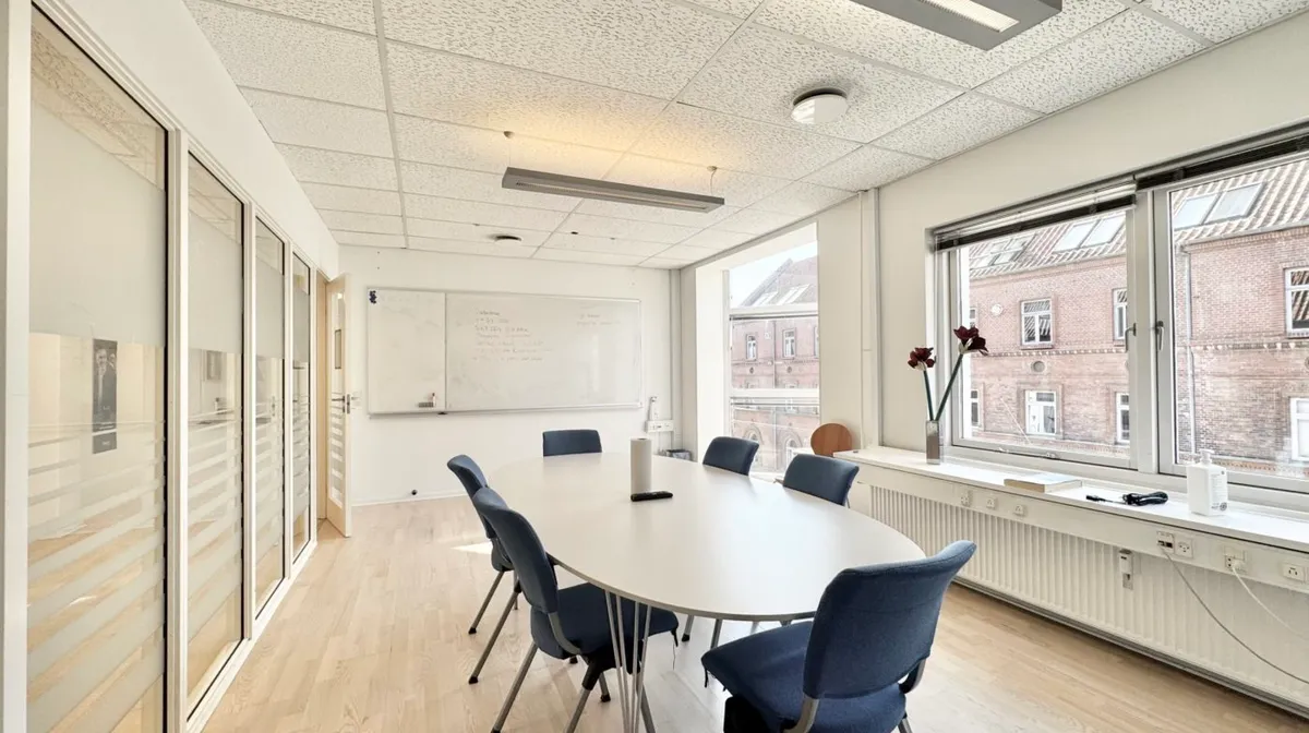 Et tomt konferencerum med hvide vægge, lyse lofter, et stort ovalt bord og blå stole samt en hvid tavle og vinduer med