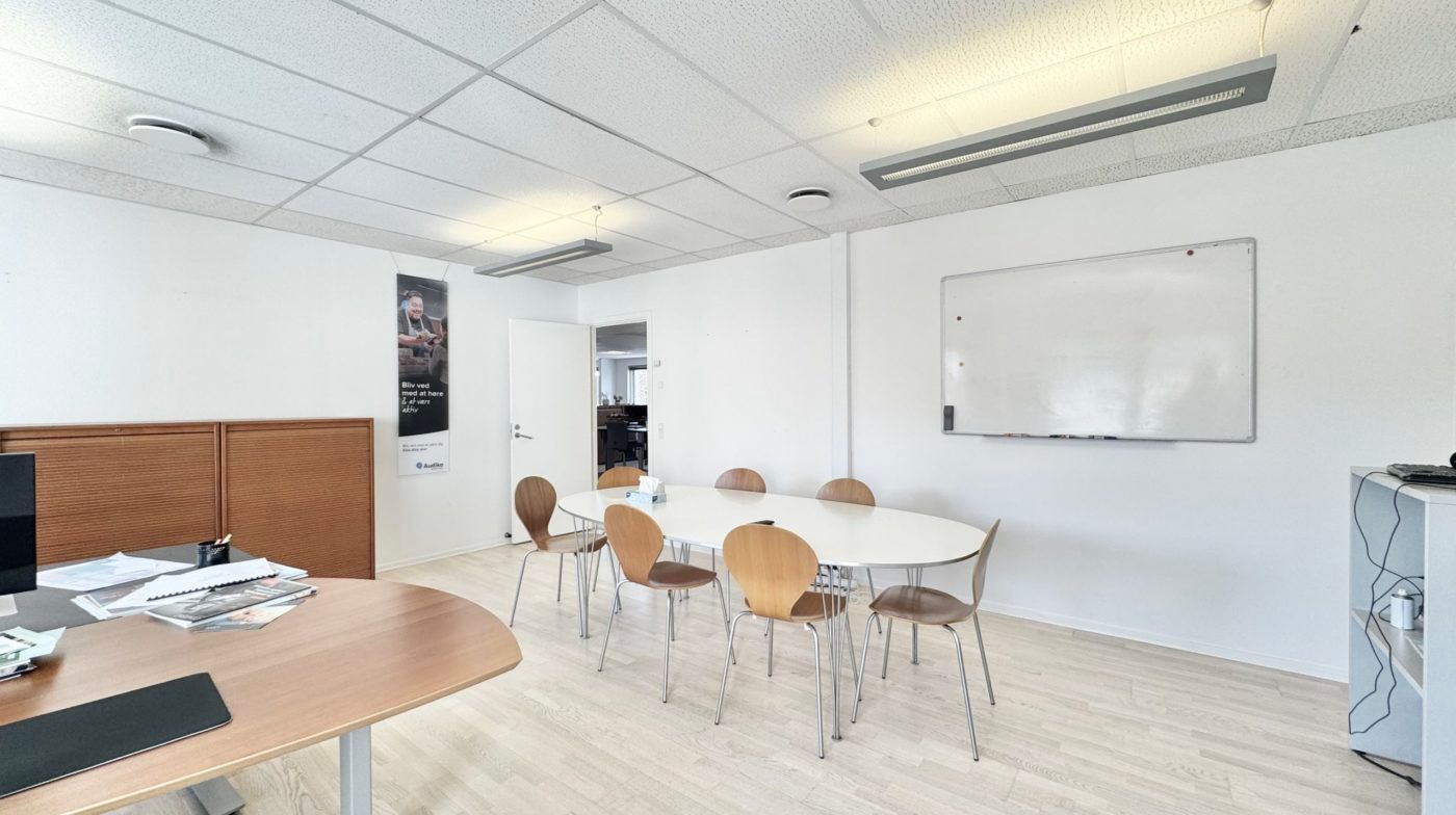 Kontor med to borde, hvor det ene bord er dækket med papirer og andre ting. Et bord med stole placeret ved en hvid væg med