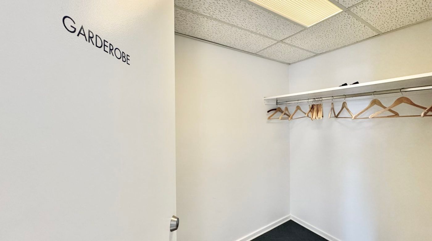 Garderobe med hængeklæder og åben dør til en anden rum. Væggene og loftet er hvide og det er mørkt på gulvet.