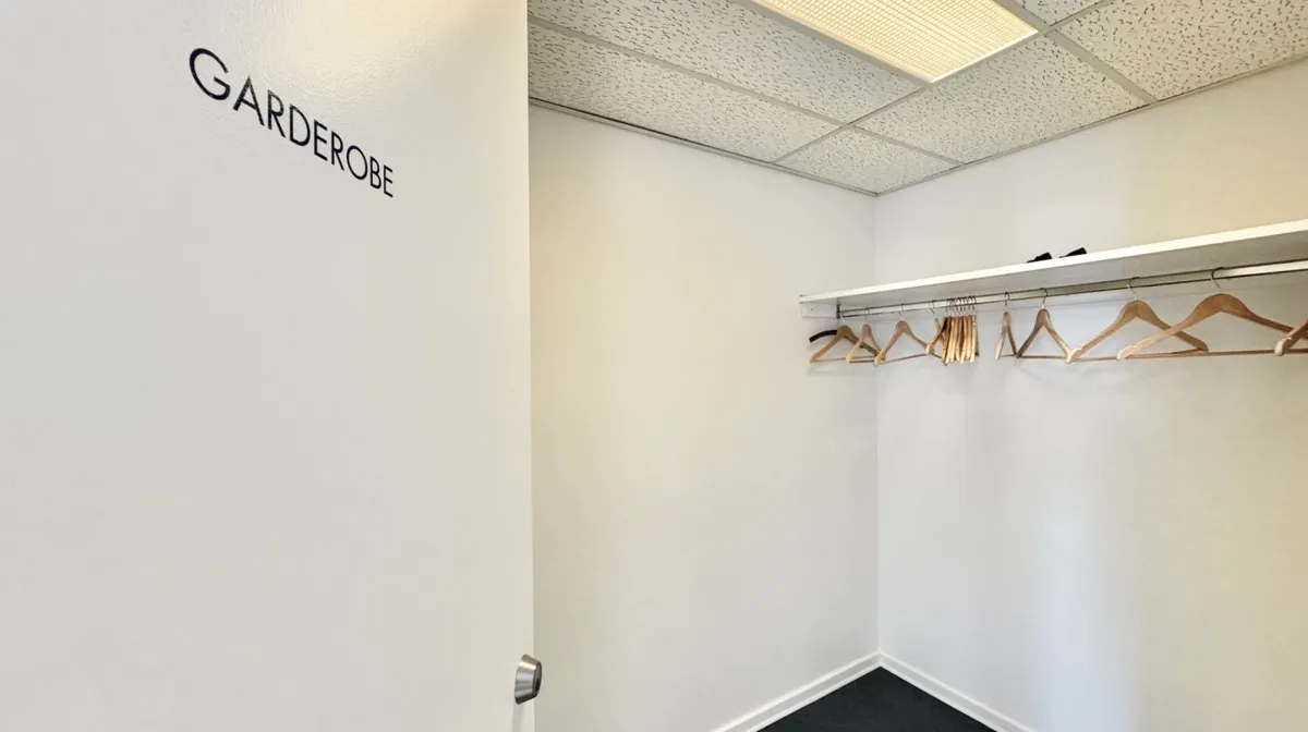 Garderobe med hængeklæder og åben dør til en anden rum. Væggene og loftet er hvide og det er mørkt på gulvet.