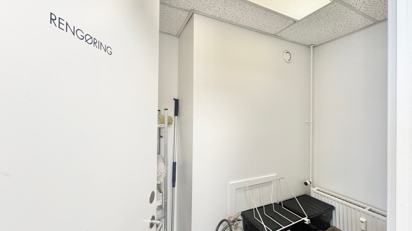 Renserirum med en hvid dør, der står åben, og hvor der er opbevaringsbokse, en stang med rengøringsredskaber og en radiator