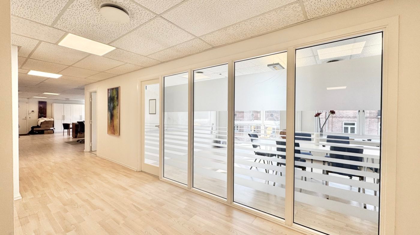 I et mødelokale er der en stor gruppe stole og et bord med en vase med blomster. Væggene er hvide, og der er vinduer med