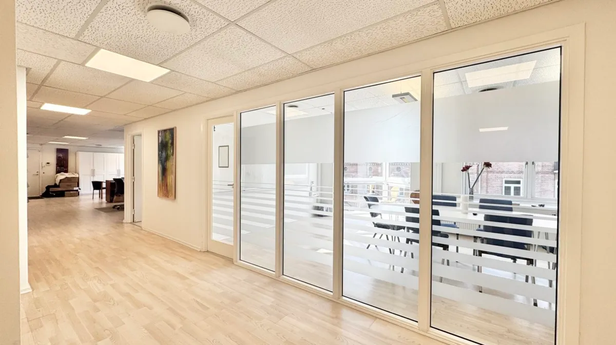 I et mødelokale er der en stor gruppe stole og et bord med en vase med blomster. Væggene er hvide, og der er vinduer med