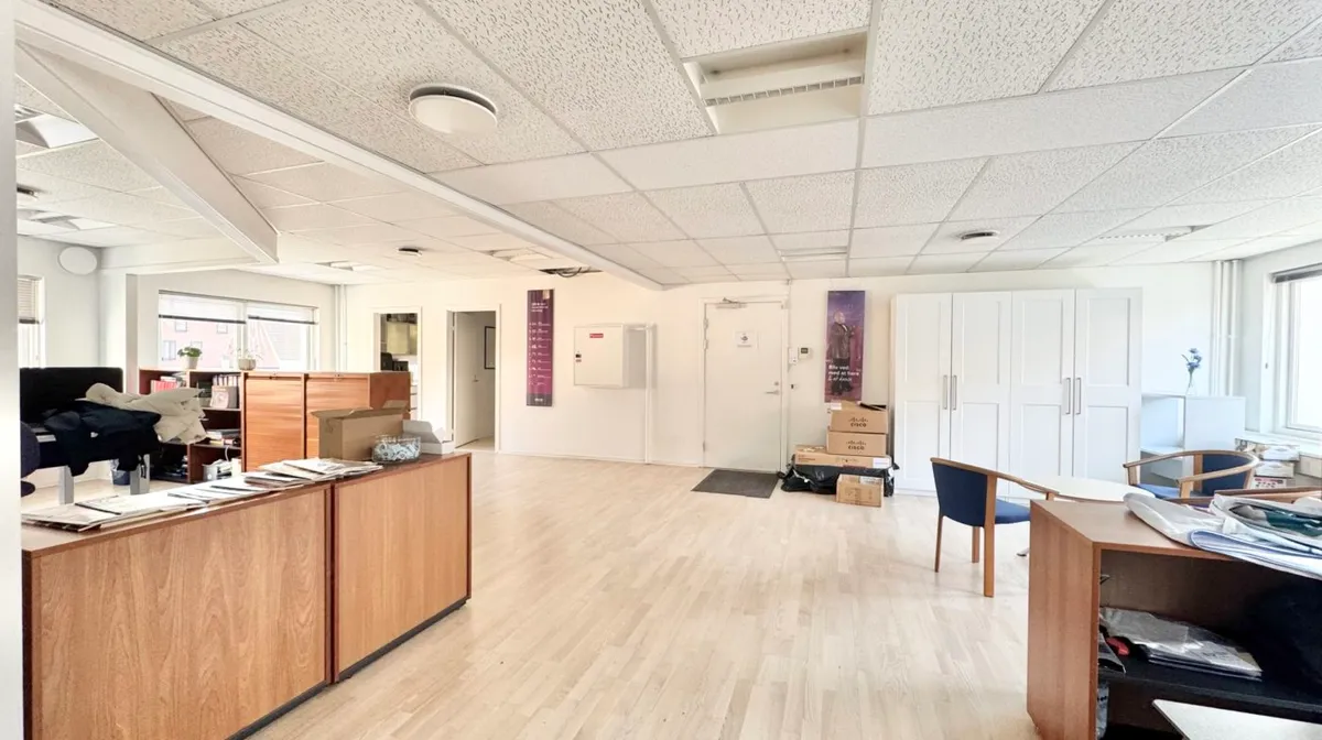 Ledig kontor i lyse og rummelige lokaler med trægulv og hvide vægge, møbler og bokse i loftet, bord og stol i hjørnet