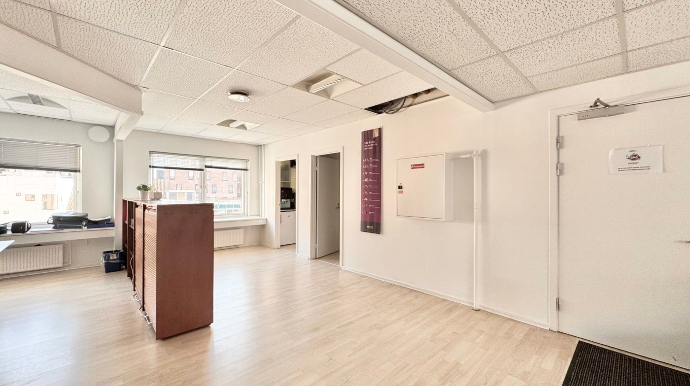 Et tomt kontor med hvid væg og trægulv, hvor der er et skab og et skrivebord med en plante og en computer.