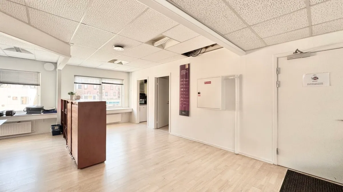 Et tomt kontor med hvid væg og trægulv, hvor der er et skab og et skrivebord med en plante og en computer.