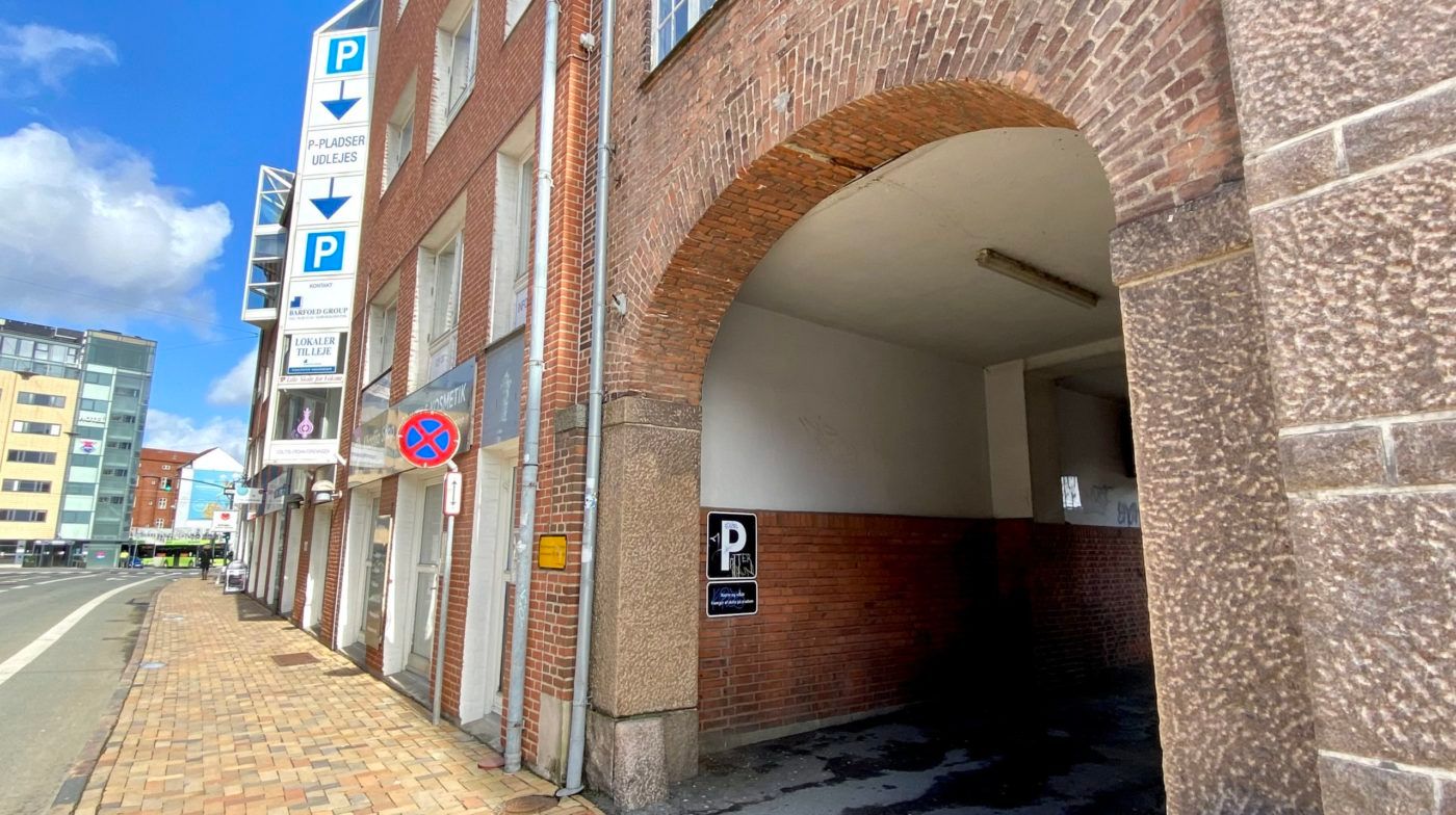 Lejlighedshus med røde mursten og borgerlige vinduer i centrum af en by. Indkørsel til parkeringsplads med skilte med rødt P