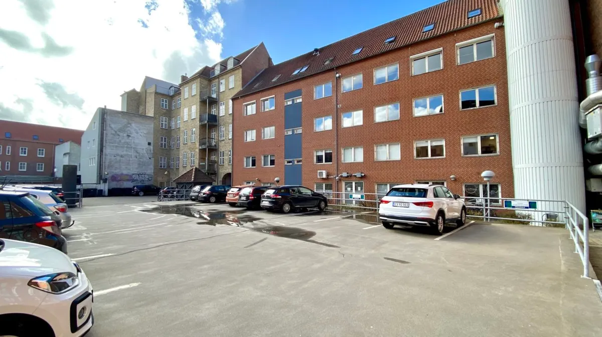 Parkeringsplads med fem biler, herunder en hvid Audi, udenfor et rødt teglstensbyggeri med flere etager og vinduer.