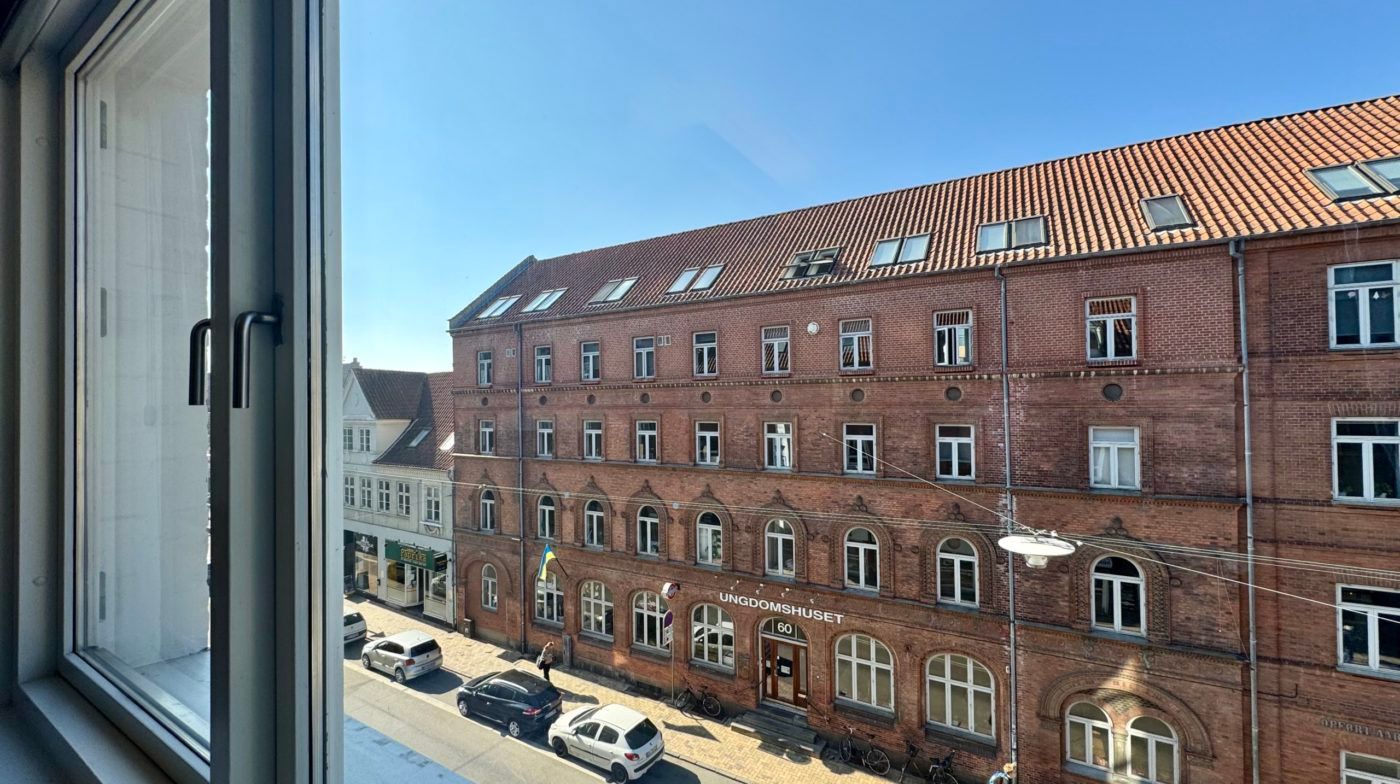 Udenfor en bygning er der to biler parkeret og en person går forbi, der er en cykel parkeret ved bygningen.