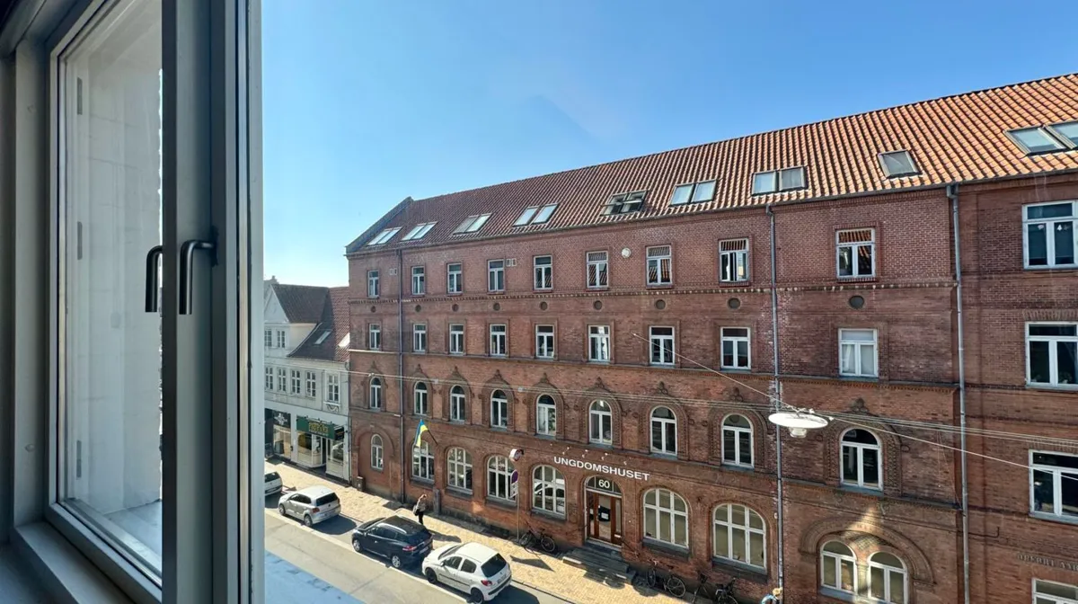 Udenfor en bygning er der to biler parkeret og en person går forbi, der er en cykel parkeret ved bygningen.