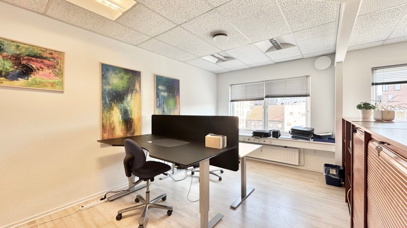 Kontor med skrivebord, stol, to malerier og et vindue med jalusi og udsigt til bygning.