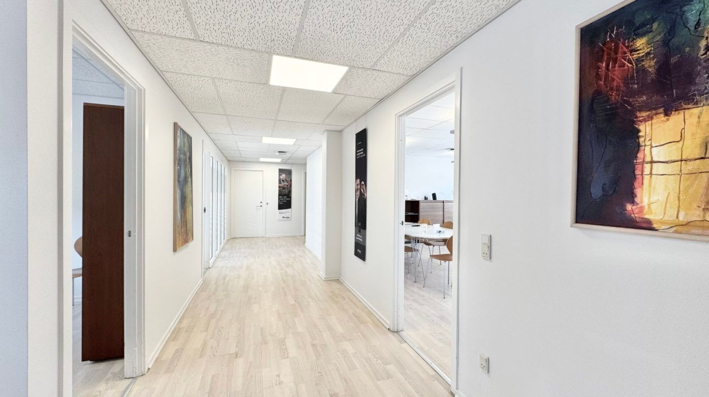 Langt og smalt kontor med lysegult gulv og hvide vægge samt kunstværker og møbler i baggrunden