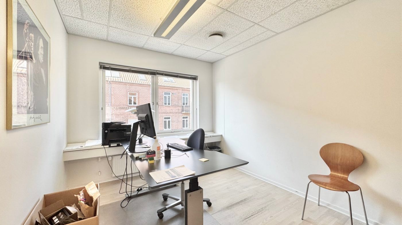 Lejlighedskontor med skrivebord, computer, stol og vinduessille med papirkurv og et maleri på væggen.