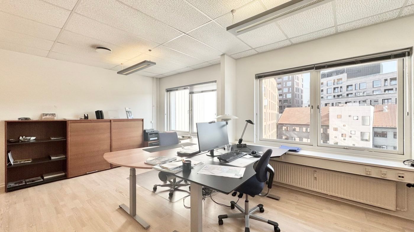 Et kontorlokale med vinduer, hvor man kan se bygninger udenfor. Et bord med en computer og en stol står mod vinduet. Et skab